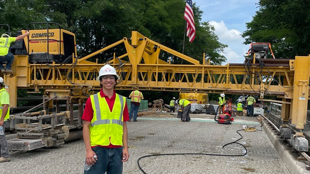 Egan Walsh in a hard hat and safety vest on a worksite in front of construction machinery.