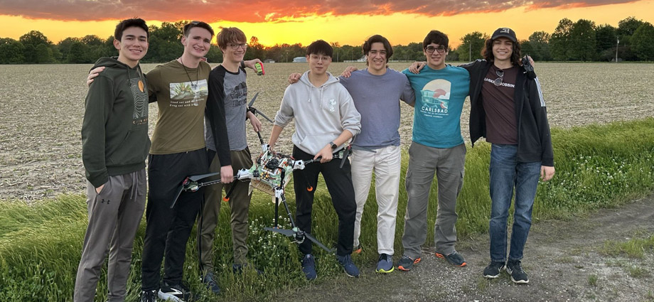 Seven students hold a drone in front of a sunset.