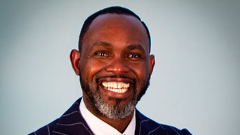 Close-up headshot of Derreck smiling with teeth showing. The top of his suit jacket can be seen.