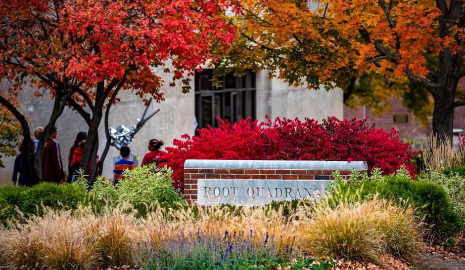 A cement and brick sign reading Root Quadrangle surrounded by colorful bushes, plants, and trees.