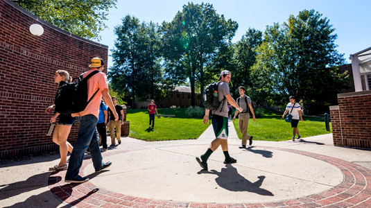 Male and female students walking on a sunny day outside an entrance to Moench Hall with the Root Quadrangle in the background.