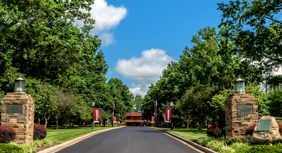 The entrance roadway into the campus of Rose-Hulman as seen from U.S. 40. Hadley Hall is visible in the distance under a blue sky.
