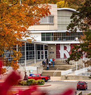 Image shows the Sports and Recreation Center through fall leaves. Students are walking up the concrete steps to the SRC entrance next to a large Rose-Hulman R.