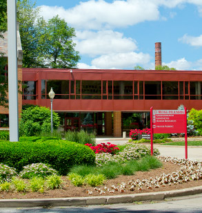 External view of the front of Hadley Hall. The campus flag pole is in the foreground amid bushes, plants and flowers.