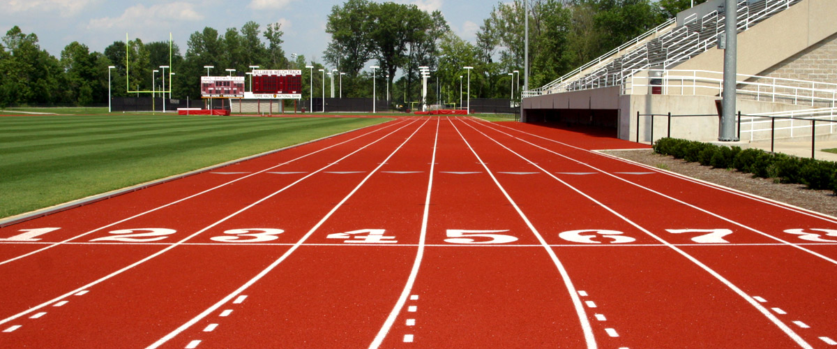 Image shows the rich, red William Welch track at Cook Stadium. The view is from just behind the eight-lane starting position just to the west of the grandstand. The green grass of the football field is to the left. 
